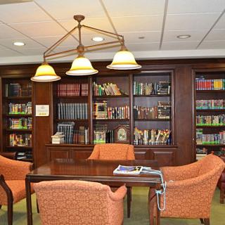A cozy library setting with a bookshelf, table, and chairs under warm lighting.