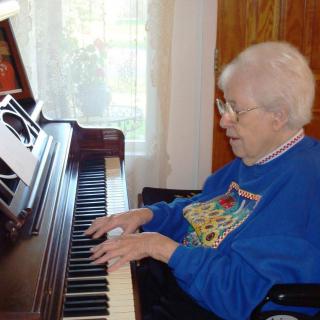 An elderly woman playing the piano indoors, wearing glasses and casual clothing.