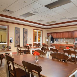 A communal dining area with wooden tables and chairs, featuring a kitchen area in the background and vases of flowers adorning the tables.