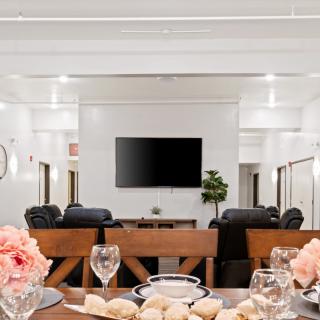 A modern living room features a dining setup with elegant place settings and floral decor, centered around a television and comfortable seating.
