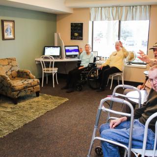 A group of seniors relax and socialize in a community room equipped with computers.