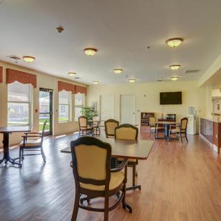 A spacious, well-lit dining area with several tables and chairs arranged on a wood floor, featuring a television mounted on the wall and large windows letting in natural light.