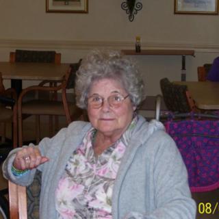 An elderly woman wearing glasses sits in a living room area.