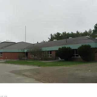 A single-story building with a sloped roof, possibly a school, surrounded by trees and overcast skies.