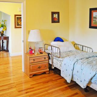 A cozy bedroom featuring a hospital bed with light blue bedding, a wooden nightstand with a lamp, and framed artwork on the walls.