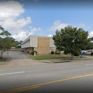 A two-story building with a parking lot and trees lining the street on a partly cloudy day.