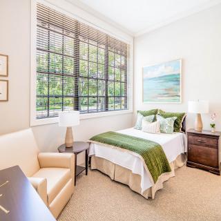 A bright and cozy bedroom with large windows, decorated with green accents and a scenic artwork.