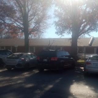 A sunny day at a parking lot with several cars and trees surrounding a building.