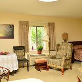 A cozy room interior with a window, various furniture including chairs, a bed, and a table adorned with a plant.