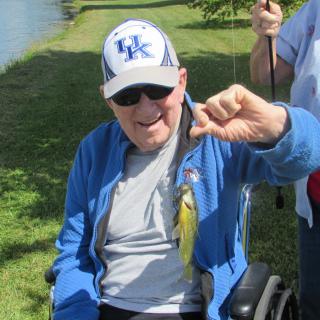 An elderly man in a wheelchair, wearing a hat and sunglasses, proudly holds up a small fish he caught by the water.
