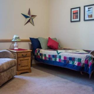 A cozy bedroom features a single bed with a colorful comforter, a few pillows, and a side table with a lamp beside an armchair. The wall is decorated with star-themed art and framed pictures.