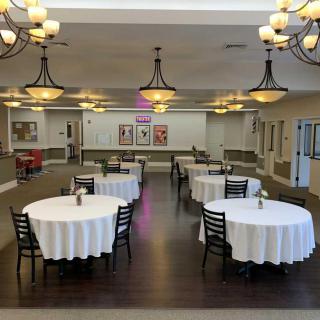 An elegant dining area with round tables covered in white tablecloths, surrounded by chairs and illuminated by chandeliers.