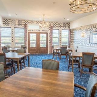 A well-lit dining area with wooden tables and chairs, featuring large windows and modern chandeliers.