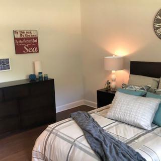 A cozy bedroom with a neatly made bed, decorative pillows, and a lamp on the nightstand. Wall art and a clock adorn the walls, creating a welcoming atmosphere.
