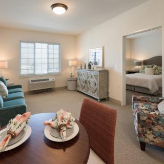 A cozy apartment featuring a living room with a sofa, dining table with place settings, and an adjacent bedroom visible through an open doorway.