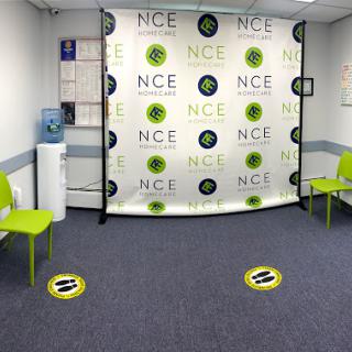 A modern waiting room featuring green chairs and a branded backdrop with social distancing markers on the floor.
