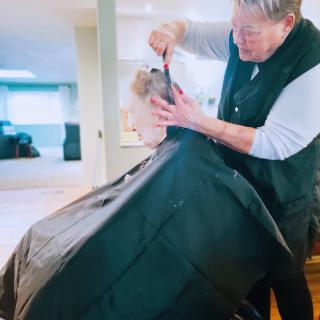 A barber is giving a haircut to a customer, covered with a black cape, in a salon setting.
