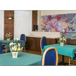 A cozy dining area with tables set up, featuring vibrant blue chairs and green tablecloths, adorned with floral centerpieces and a colorful mural on the wall.