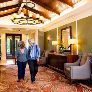 Two people walk through a well-decorated hallway with comfortable seating and warm lighting.