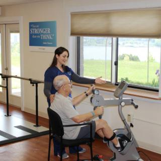 An elderly man receives guidance from a healthcare professional while using exercise equipment.