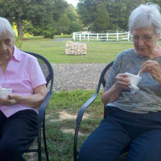 Two elderly women enjoying ice cream while sitting outside in a garden setting.