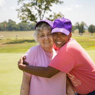 Two people hugging and smiling outdoors on a sunny day, wearing hats.