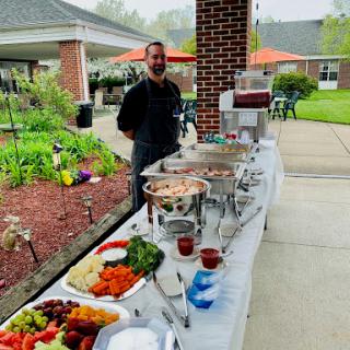A chef stands proudly behind a buffet setup featuring a variety of fresh vegetables and dishes in an outdoor garden setting.