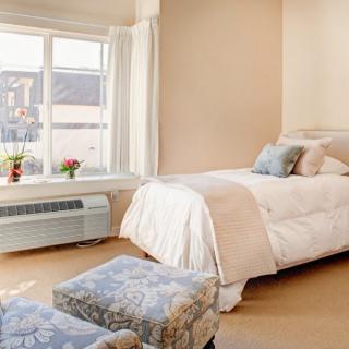 A cozy bedroom featuring a single bed with light-colored bedding, soft cushions, and a large window allowing natural light to fill the space.