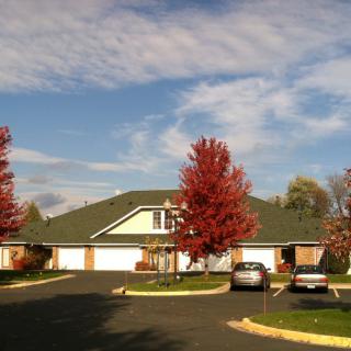 A suburban house with autumn trees and cars parked in the driveway under a partly cloudy sky.