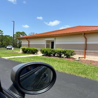 A view of a modern building with a red roof, captured from the side mirror of a car on a sunny day.