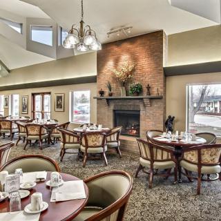 A cozy restaurant dining area featuring round tables, elegant chairs, and a warm fireplace surrounded by large windows.