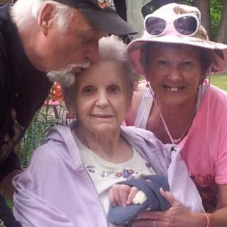 An elderly woman is surrounded by loved ones, enjoying a moment together outdoors.