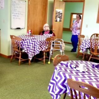 An elderly person sits in a dining room with zigzag-patterned tablecloths, accompanied by a caregiver.
