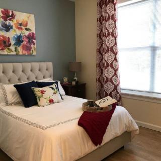 A cozy bedroom with a neatly made bed, featuring floral artwork and decorative pillows, complemented by patterned curtains and a nightstand with a lamp.