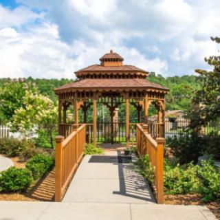A scenic view of a wooden gazebo connected by a bridge in a lush garden setting.