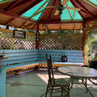 A cozy gazebo with a blue bench and round table, featuring a ceiling fan and lattice walls.