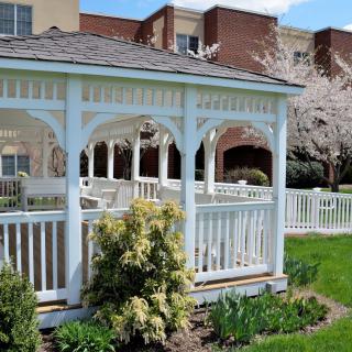 A charming white gazebo surrounded by greenery and a building in the background, capturing a serene springtime scene.