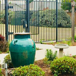 A tranquil garden scene featuring a large green vase and a birdbath amidst neatly trimmed shrubs, with a tall fence in the background.