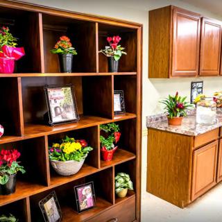 An indoor setting featuring wooden shelves with potted plants and flowers beside kitchen cabinets.