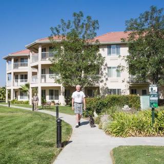 A man walks along a sidewalk in a well-maintained retirement community with landscaped grounds and a multi-story residential building.