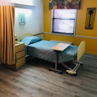 A cozy hospital room featuring a neatly made bed, wooden furniture, and warm yellow walls.