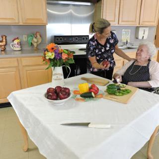 A caregiver assists an elderly person in a kitchen filled with fresh fruits and vegetables.