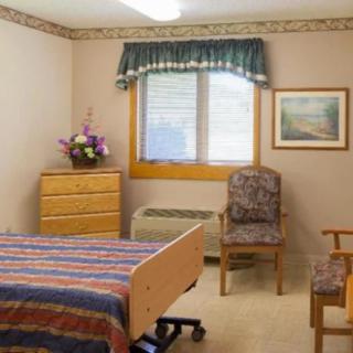 A hospital room with a bed, dresser, and chairs near a window adorned with curtains and a flower vase.