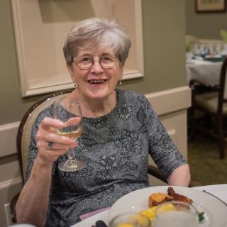 An elderly woman enjoys a meal at a restaurant, raising her wine glass with a smile.
