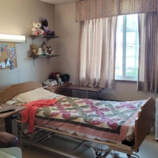 A cozy hospital room with a bed covered in a quilt, shelves adorned with toys, and light filtering through the curtains.
