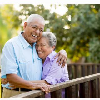 An elderly couple embraces warmly on a sunny day outdoors.