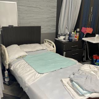 A simple bedroom setting featuring a hospital-style bed with fresh bedding, a nightstand filled with personal items, and draped curtains.