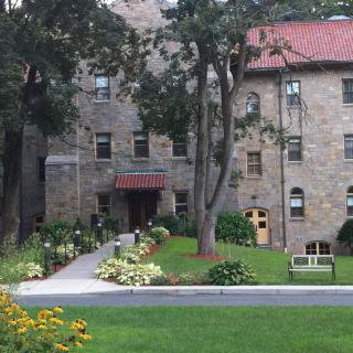 A stone building surrounded by lush landscaping and trees, with a walkway leading to the entrance.