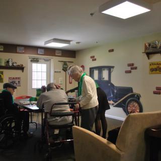 A group of elderly individuals engaged in an activity in a communal room decorated with vintage car images.