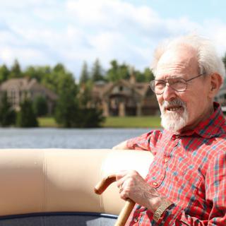 An elderly man sitting on a boat near a lake, holding a cane on a sunny day.